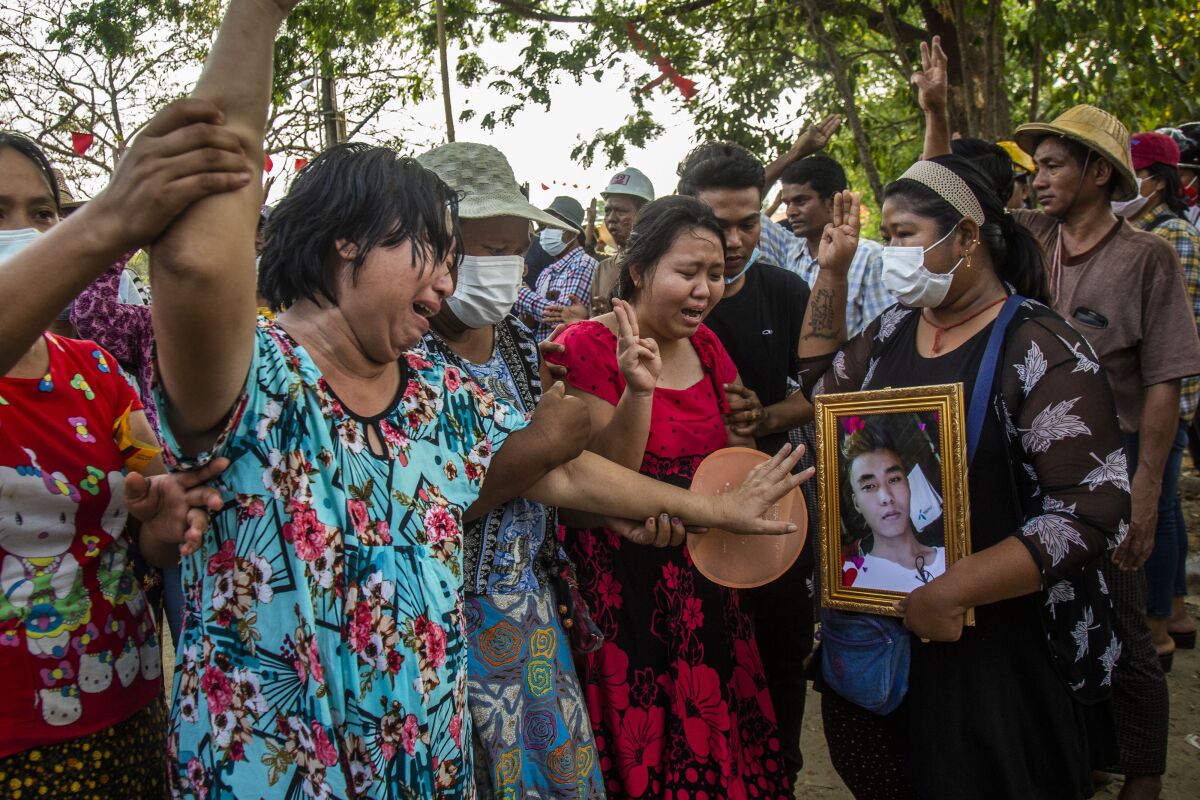 Women in colorful dresses hold up three fingers as they weep next to a woman carrying a framed photo of a man