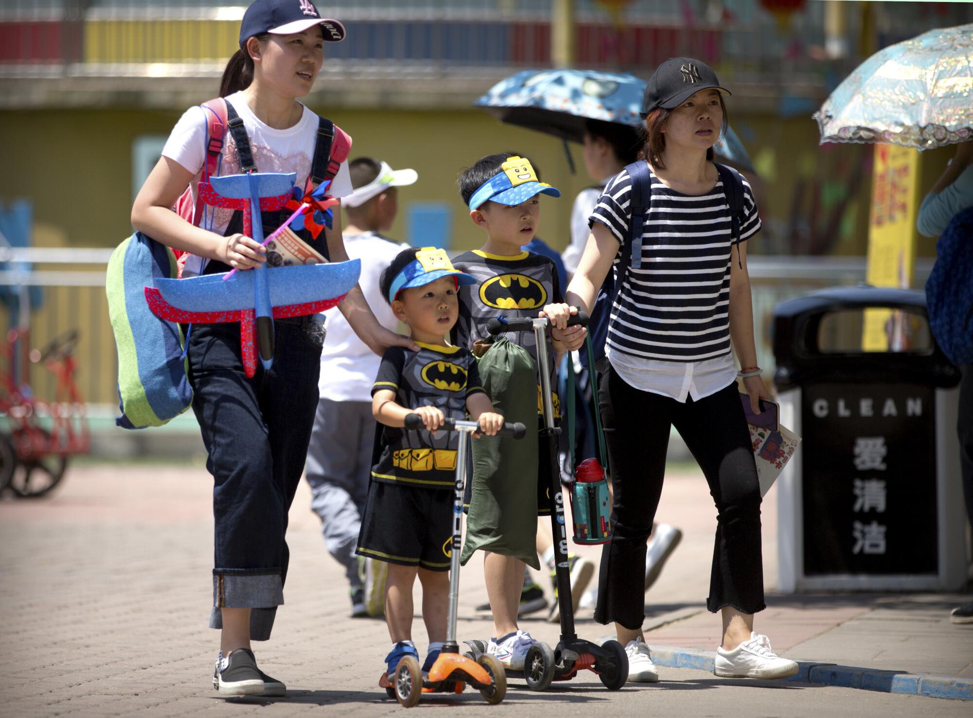 Duas mulheres, uma delas carregando brinquedos, caminham com crianças em roupas combinando e em patinetes em um parque público 