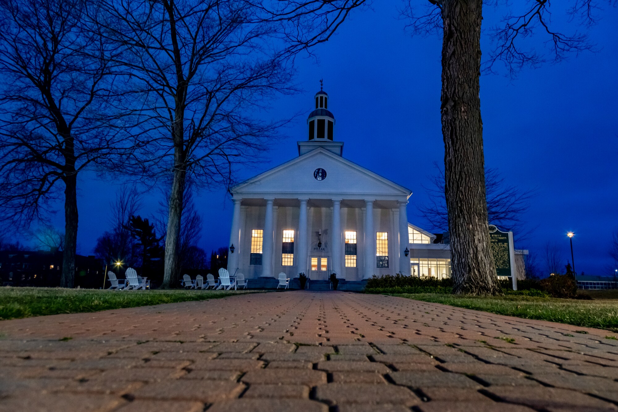 Light shines from a white building with several columns, sitting by bare trees against a dark sky at the end of a brick path.