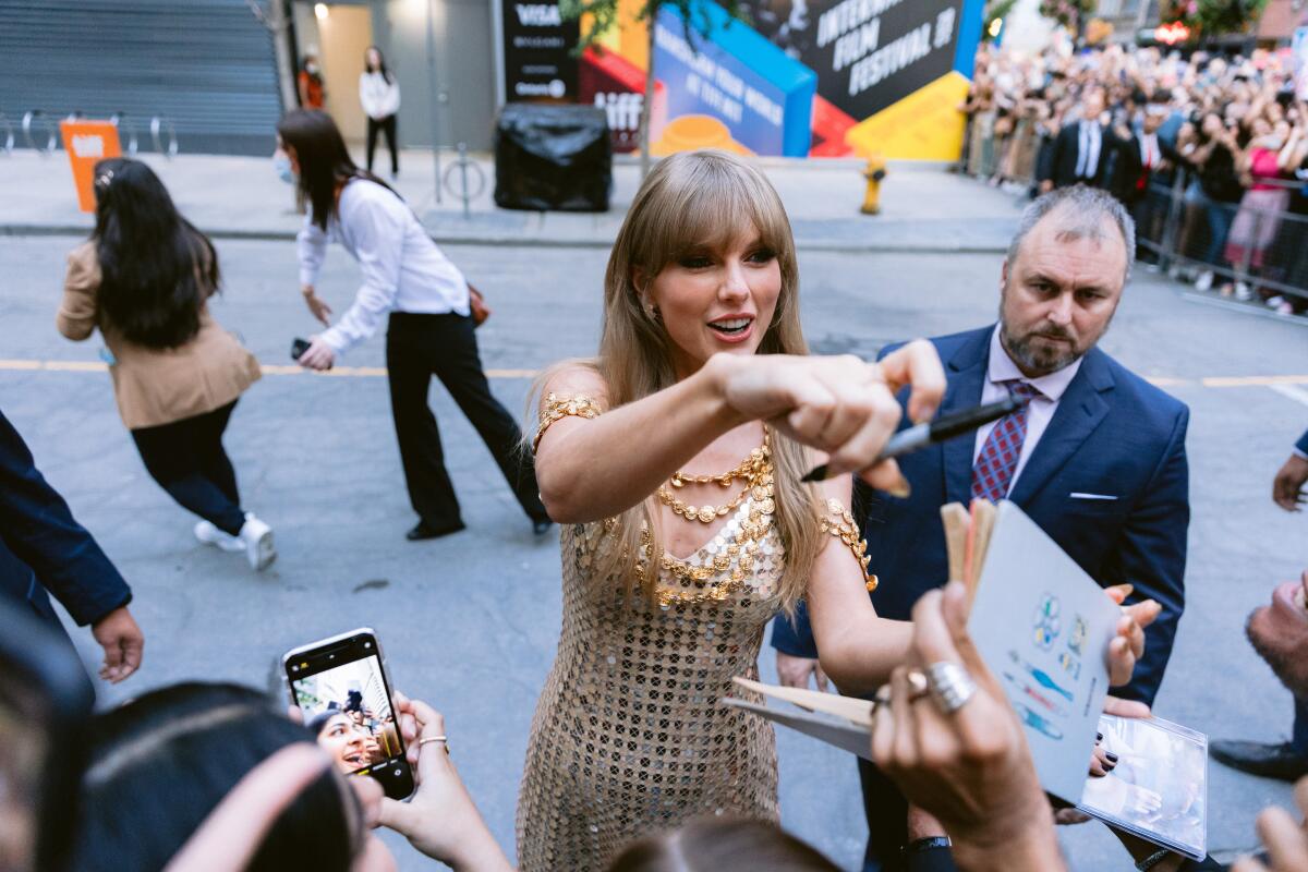 Taylor Swift, wearing a gold sparkly dress,  greets fans on a sidewalk.