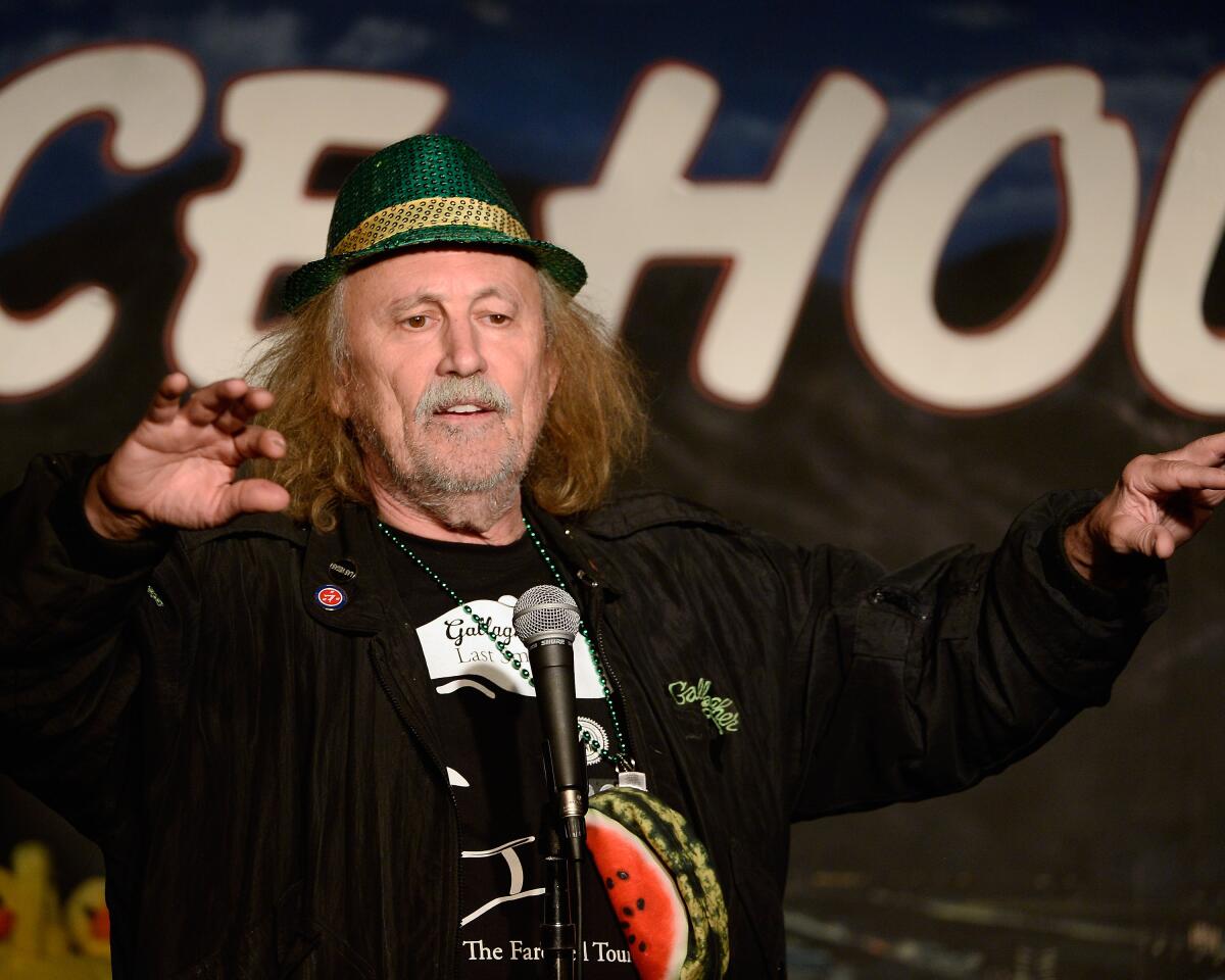 A man with long hair and wearing a green hat gestures in front of a microphone