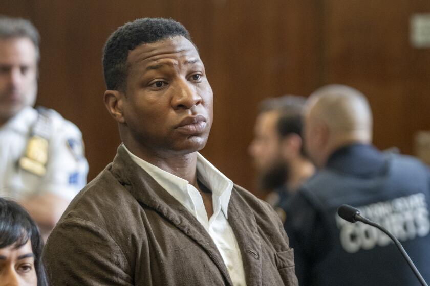 Jonathan Majors is standing in a courtroom with a serious face while wearing a brown coat and white shirt.