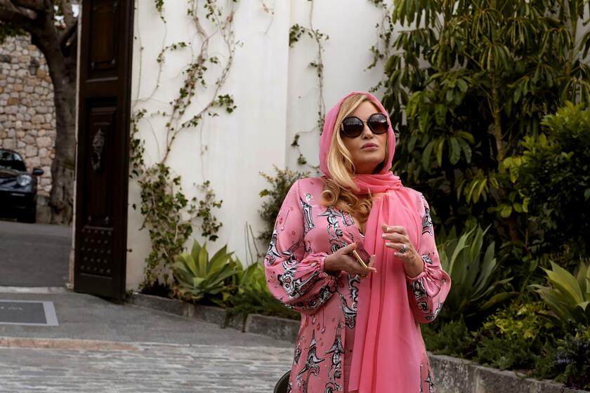 A woman in a pink head scarf, dress and sunglasses stand by an olive green Vespa.