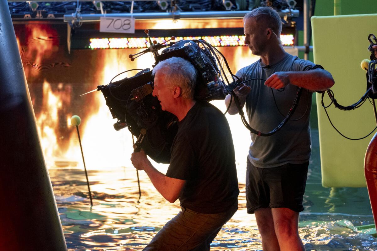 Director James Cameron stands in water holding a giant movie camera on his shoulder