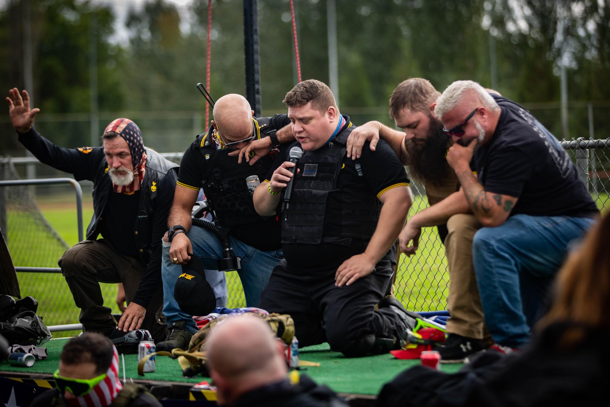 A man in a tactical vest speaks into a microphone among group of men kneeling on stage