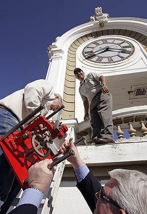 Tim Rush, right, of the Santa Ana Historical Society helps antique clock expert Larry Squires haul the restored gearbox to the Spurgeon Building clock tower. Waiting on the rooftop tower is Jesse Alvidrez from the S.A. Community Development Agency. The 1913, four-sided timepiece should now keep correct time.
