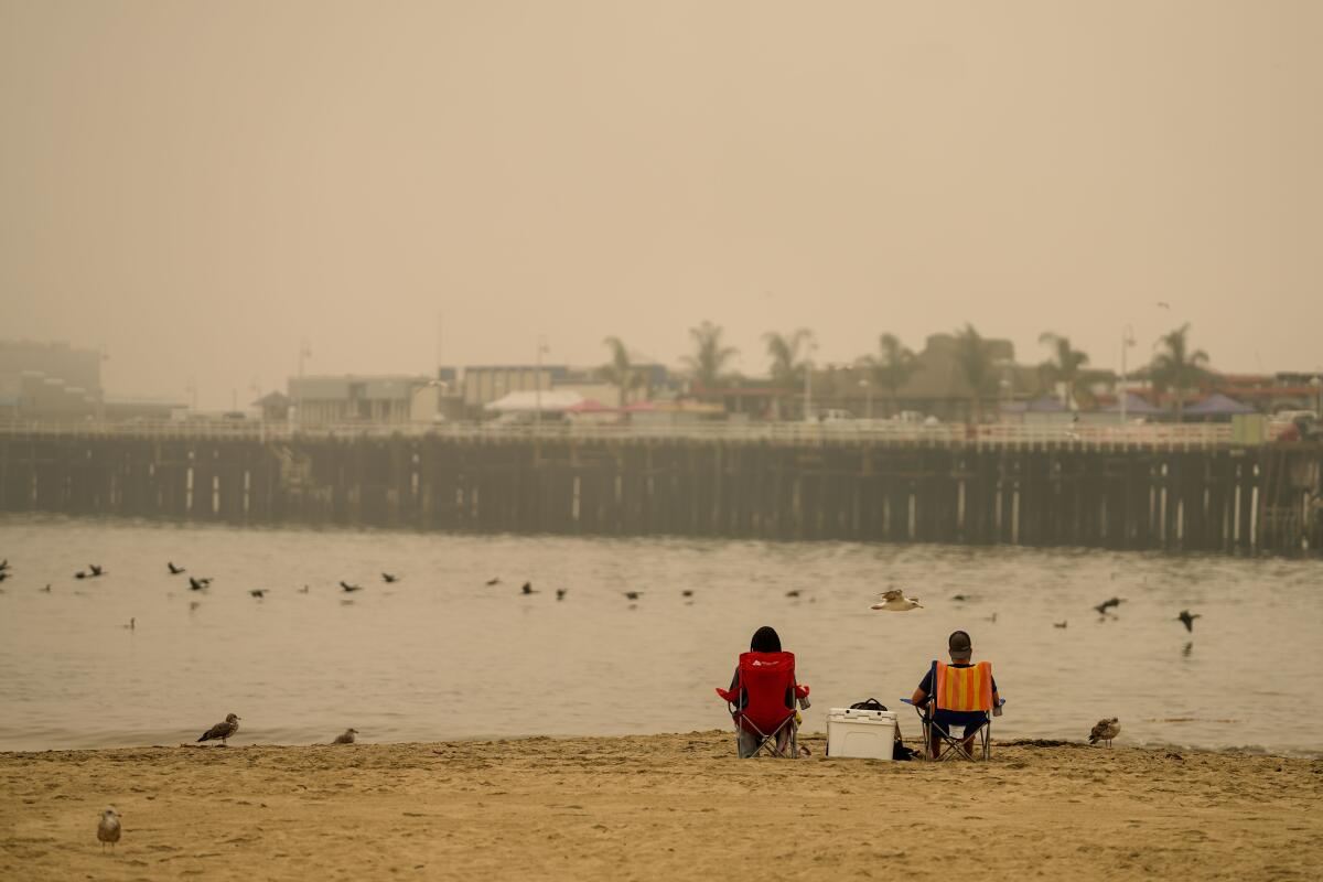 People sit on an empty beach in Santa Cruz as smoke from the wildfires obscures the view of the Santa Cruz Wharf.