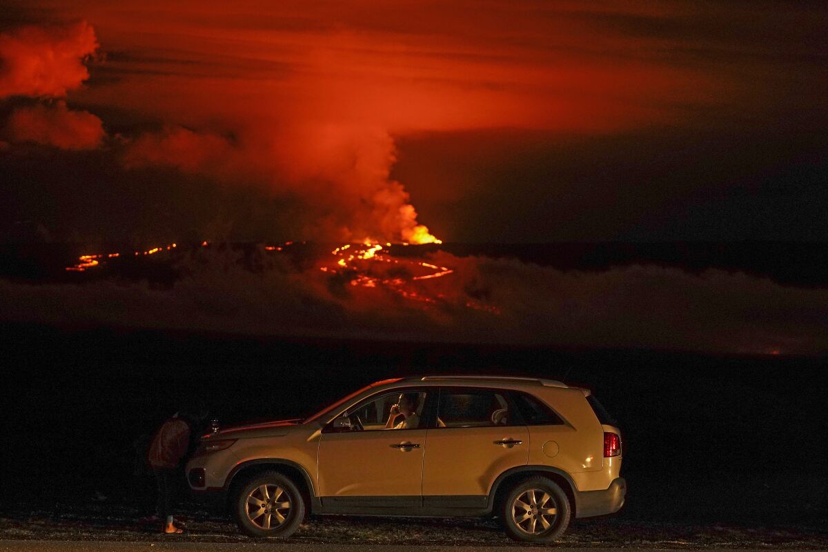 Mauna Loa can be seen erupting behind a car in which a man is talking on his phone.