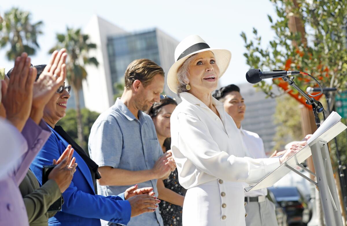 A woman in white blouse and pants and white hat speaks into a microphone as people standing behind her applaud.