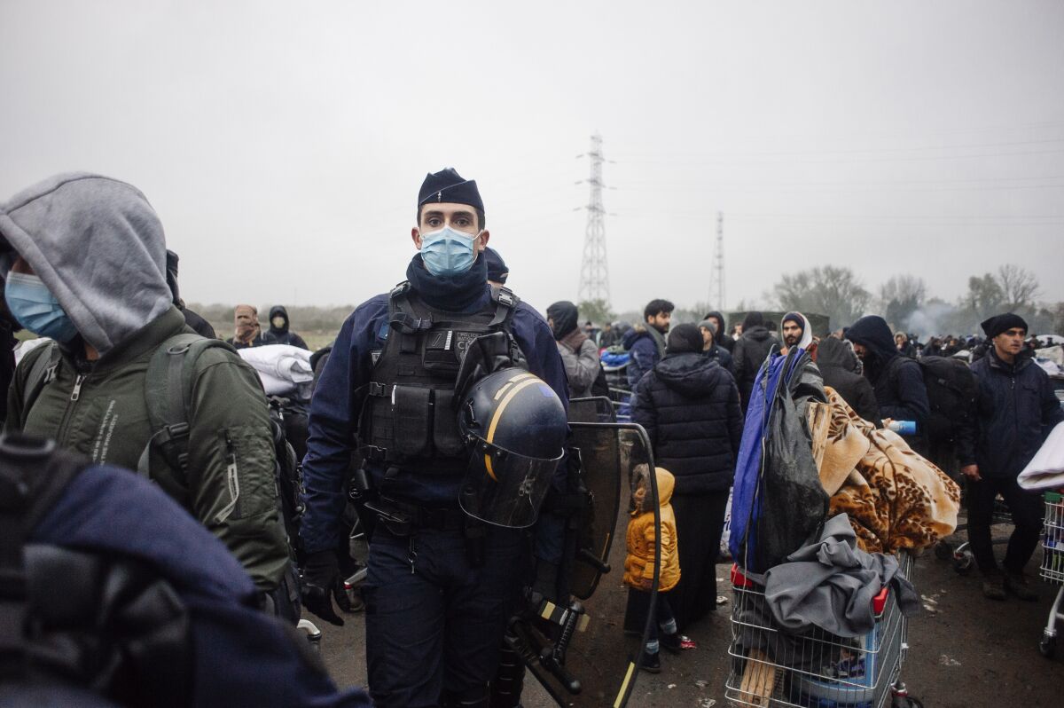 A police officer looks on as migrants gather in a camp in Grande-Synthe, Northern France, Tuesday, Nov. 16, 2021. French police were evacuating migrants from a makeshift camp near Dunkirk, in northern France, where at least 1,500 people gathered in hopes of making it across the English Channel to Britain. Migrants, including some families with young children, could be seen packing their few belongings as police were encircling the camp, on the site of a former industrial complex in Grande-Synthe, east of Dunkirk. (AP Photo/Louis Witter)