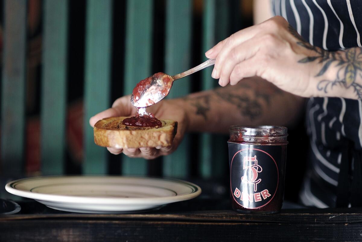A hand spoons strawberry jam onto a piece of bread. In front is a glass jar of the jam.