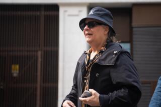 Duran Duran's Andy Taylor walks and smiles while wearing a black bucket hat and jacket  and a brown and  black shirt