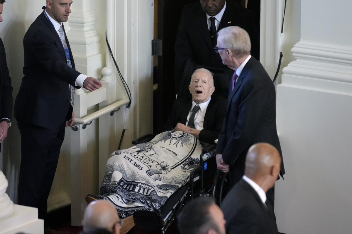 Former President Carter is wheeled into a church, a blanket over his knees.