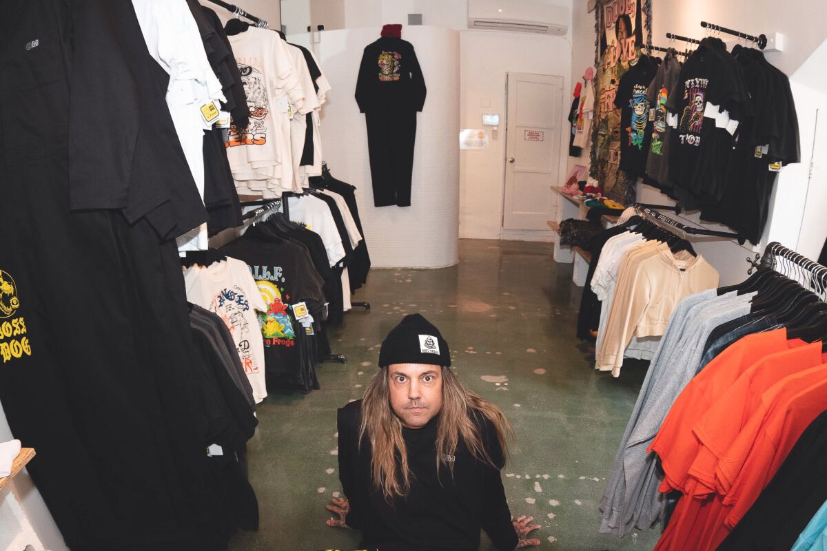 A person with long hair in a knit hat crouches among racks of clothing in a store.