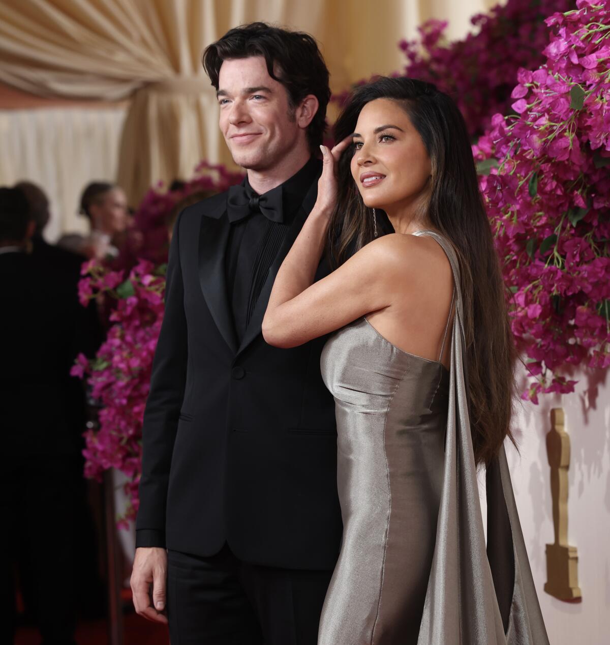 John Mulaney in a black suit posing next to Olivia Munn in a silver gown in front of hanging flowers