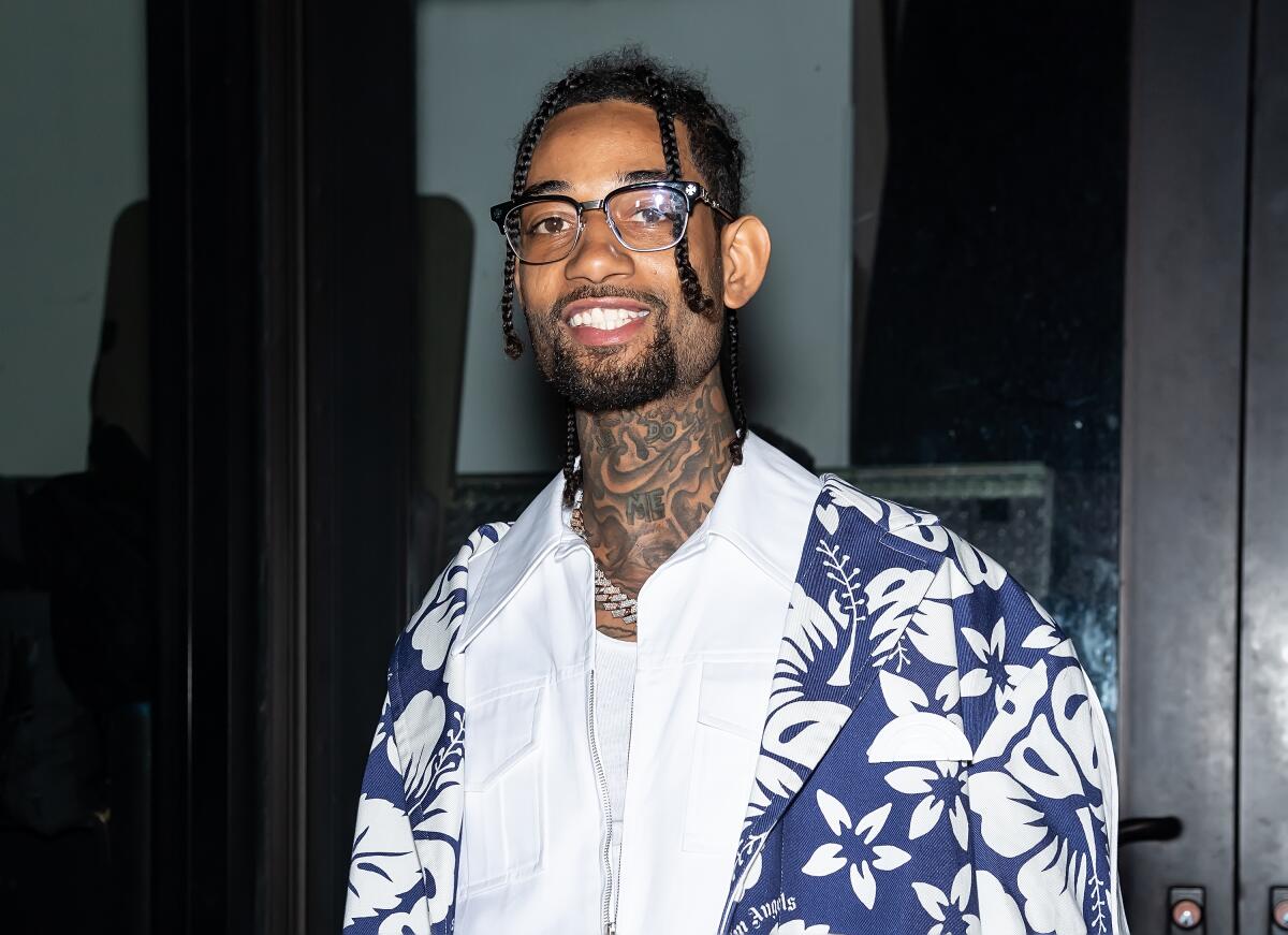 PnB Rock smiles while wearing glasses, a chain and a blue and white Hawaiian shirt 