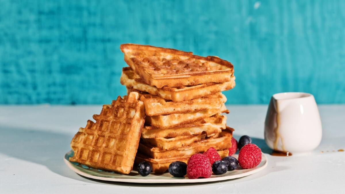 A stack of golden waffles, cut into triangles, on a plate with raspberries and blueberries, next to a container of syrup.