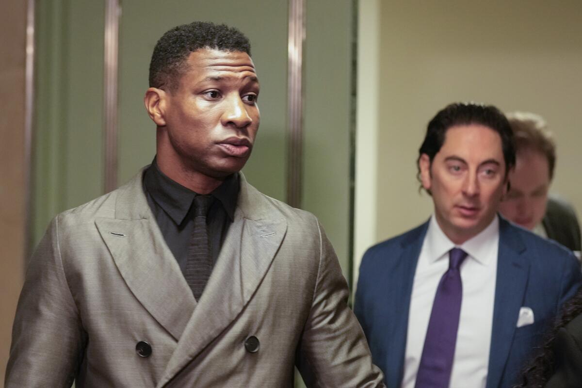 Jonathan Majors in front of a door in a gray suit in front of a man in a blue suit jacket and tie