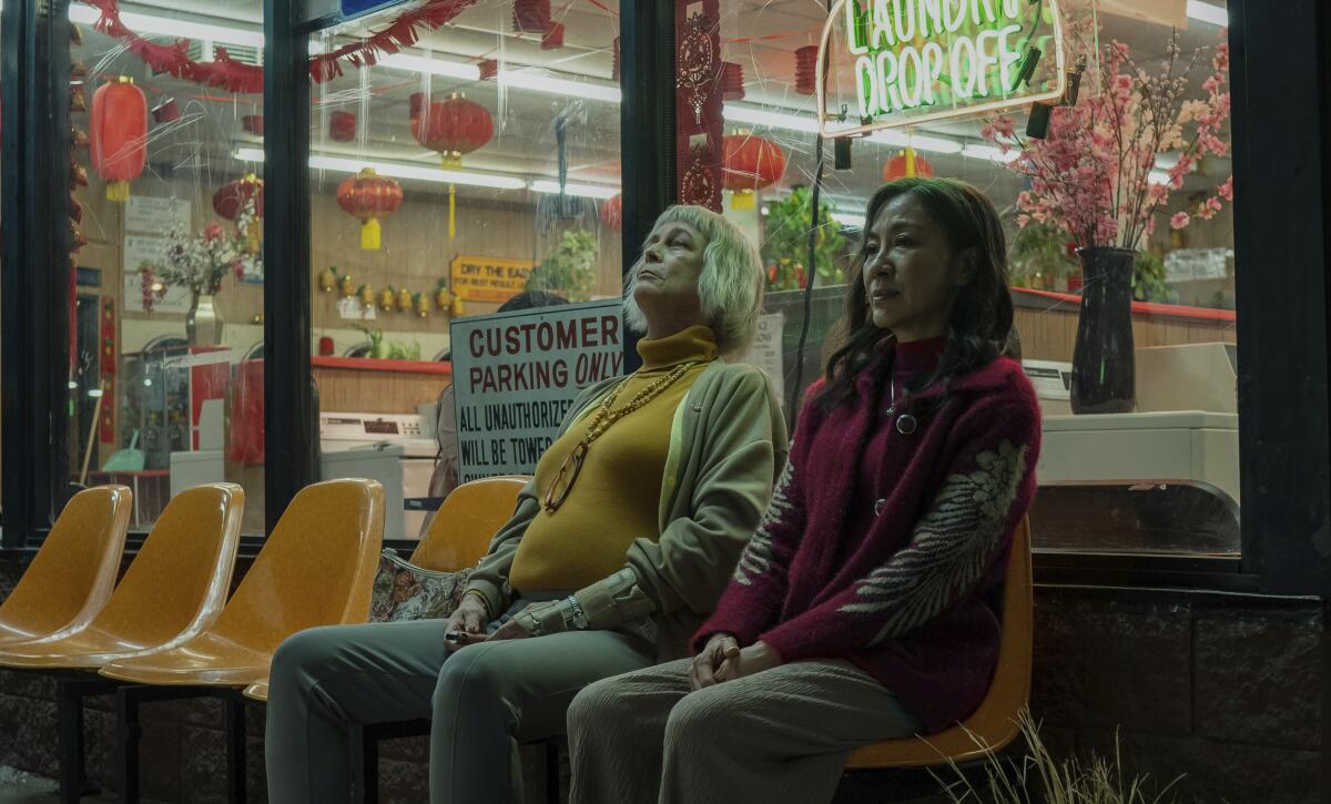 Two women sit side by side in molded plastic chairs outside a laundromat's glass window.