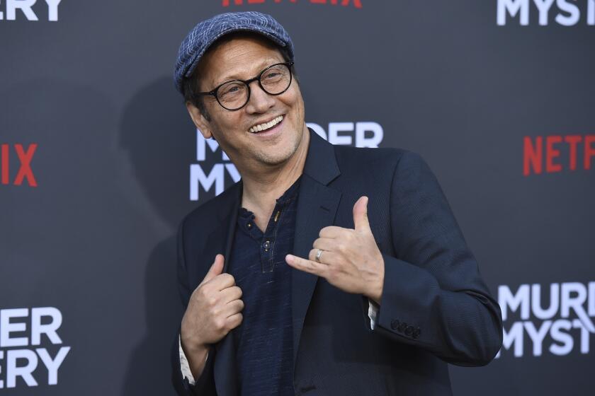 Rob Schneider, wearing a dark suit, flashes a Shaka sign at a red carpet