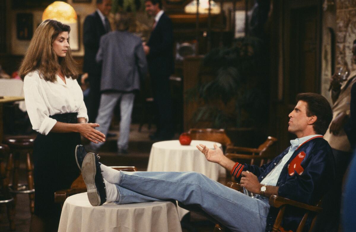 A female bartender talks to a man with his feet up on a table.