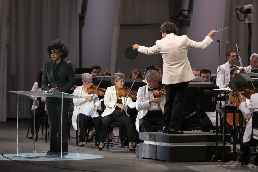 A man in a white jacket conducts members of an orchestra, also wearing white jackets, while a woman in black is at a podium.