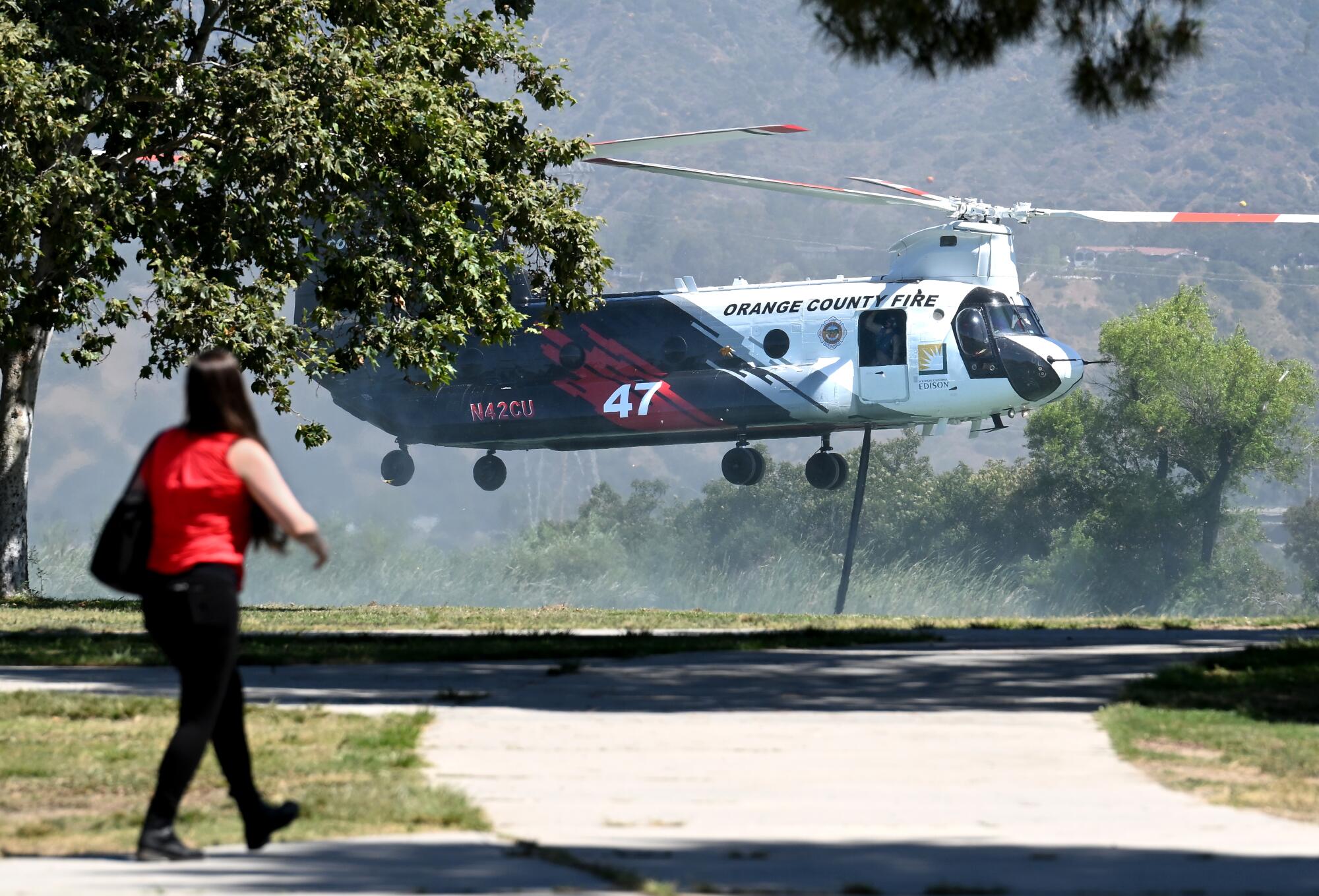 A firefighting helicopter hovers just above ground as a figure walks in the foreground.