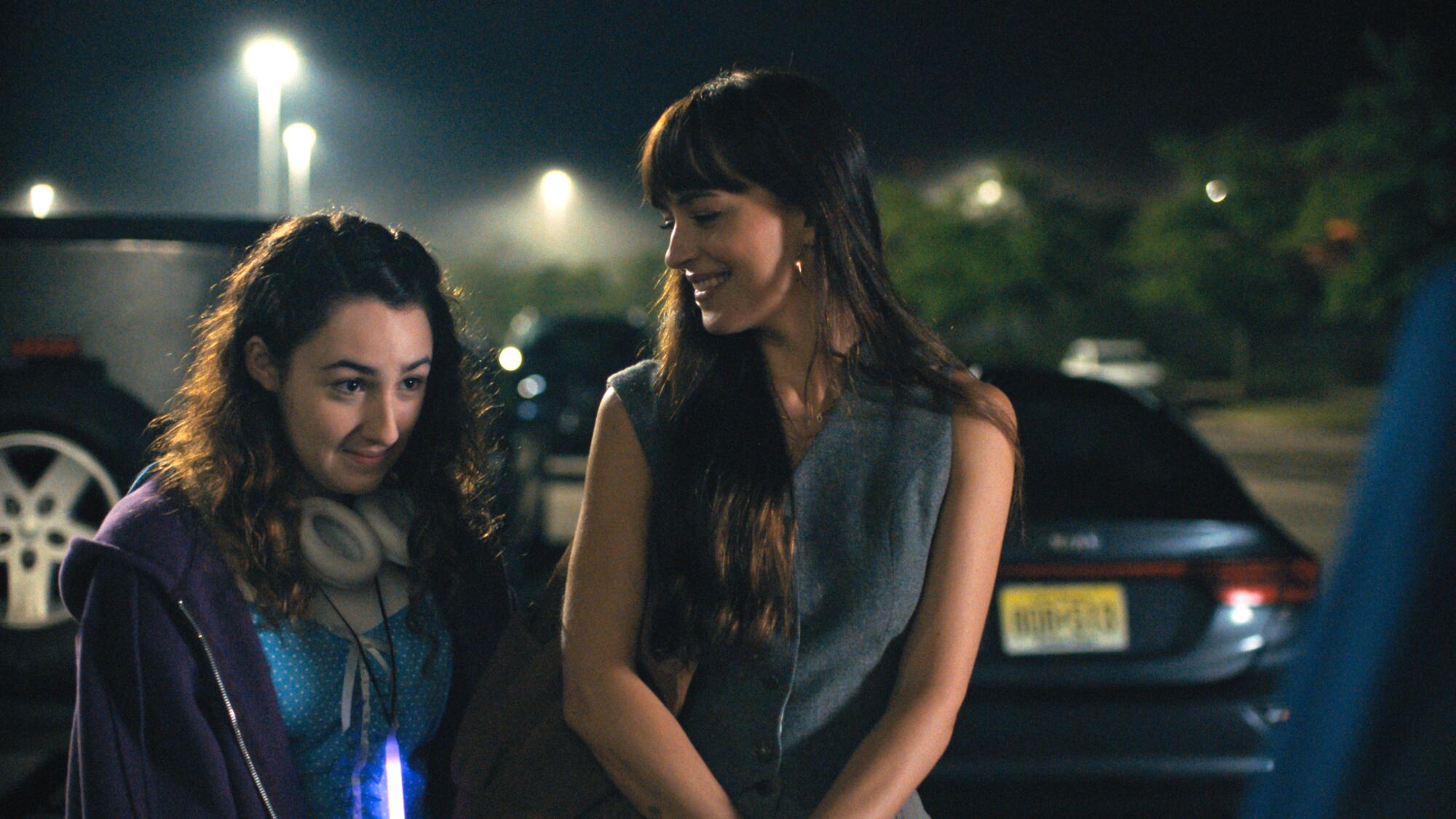 A woman smiles at a younger woman next to her in a parking lot.