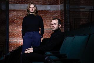 Micah Gottlieb, right, sits in a chair, and Sarah Winshall, stands to his left in front of a brick wall. 