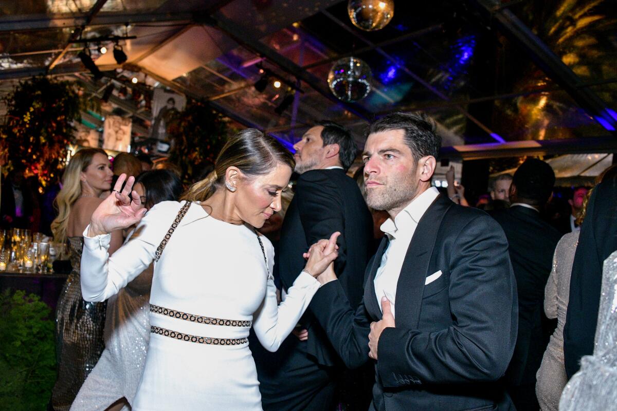 A woman in a white dress and a man in a white button-down and suit jacket hold hands on a dance floor.