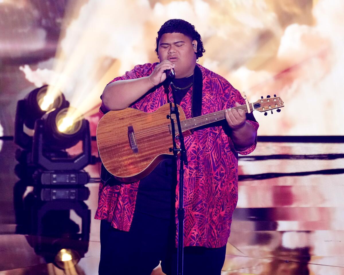 Iam Tongi closes his eyes while singing and playing guitar in front of a microphone onstage.