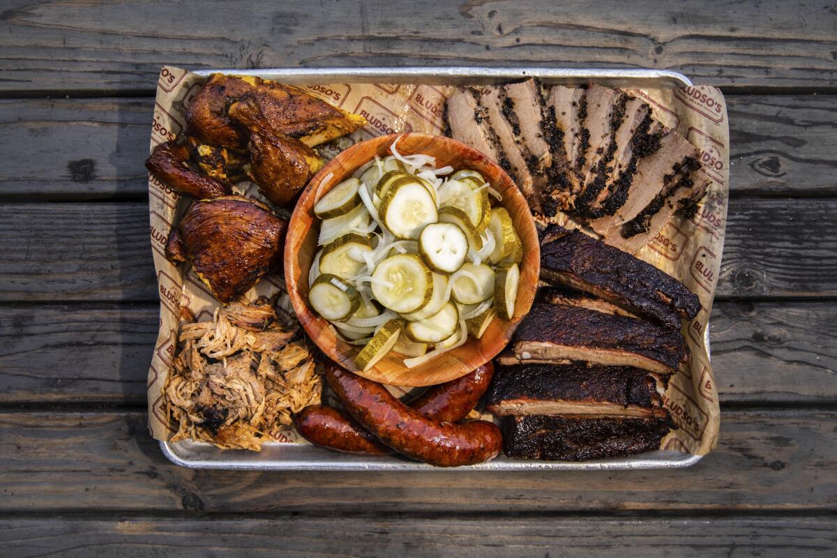 A tray with a bowl of homemade pickles in the center surrounded by barbecued chicken, brisket, pork ribs and two sausages