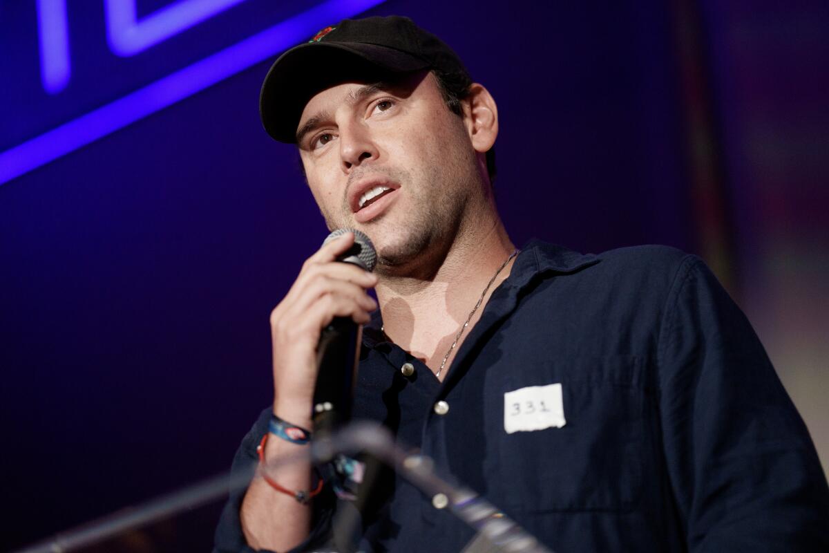 A man in a dark shirt and dark ballcap holds a mic while speaking