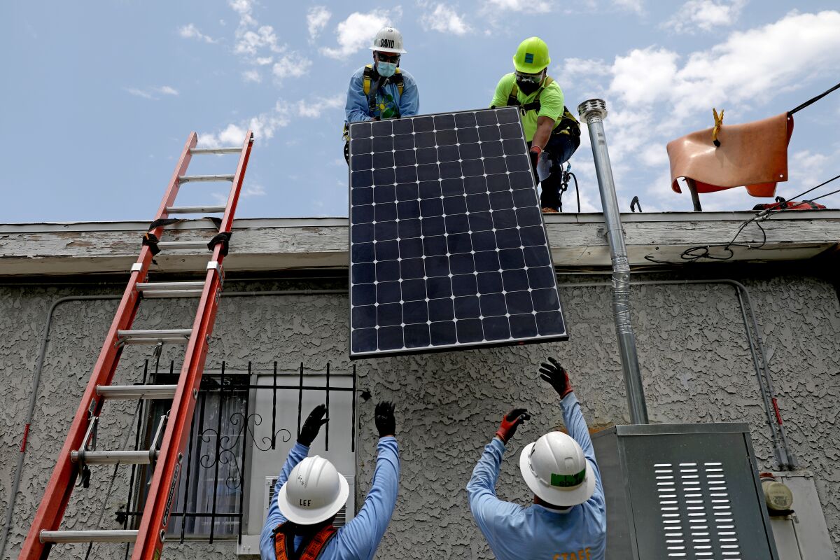 Two workers on the ground hand a solar panel up to two workers on a roof 