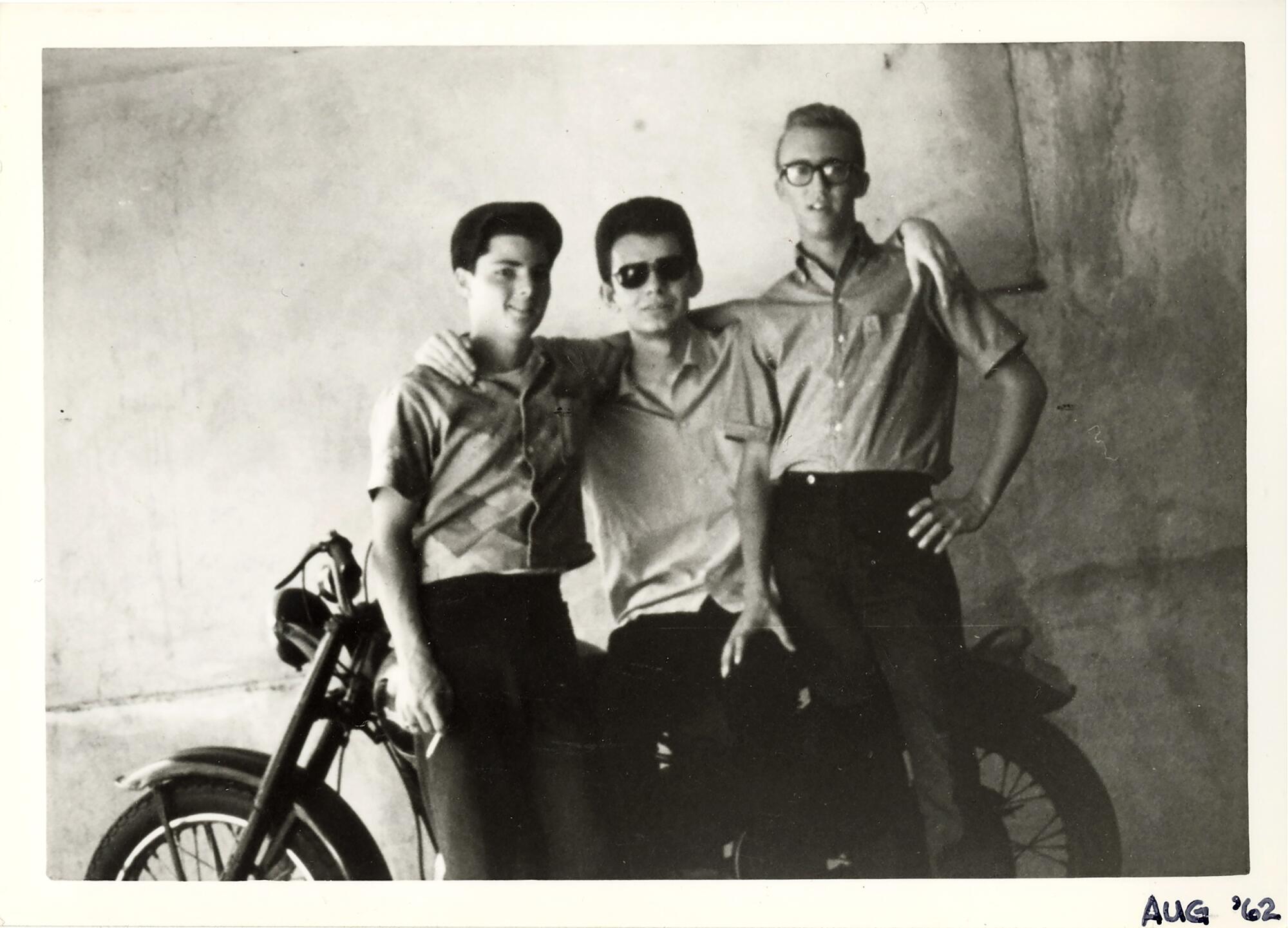 Three men stand in front of a motorcycle.