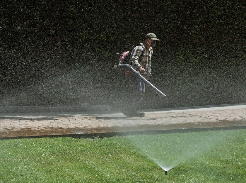 Un jardinero usa un soplador de hojas mientras limpia un camino de entrada mientras los rociadores riegan el césped verde de una casa de Los Ángeles. 