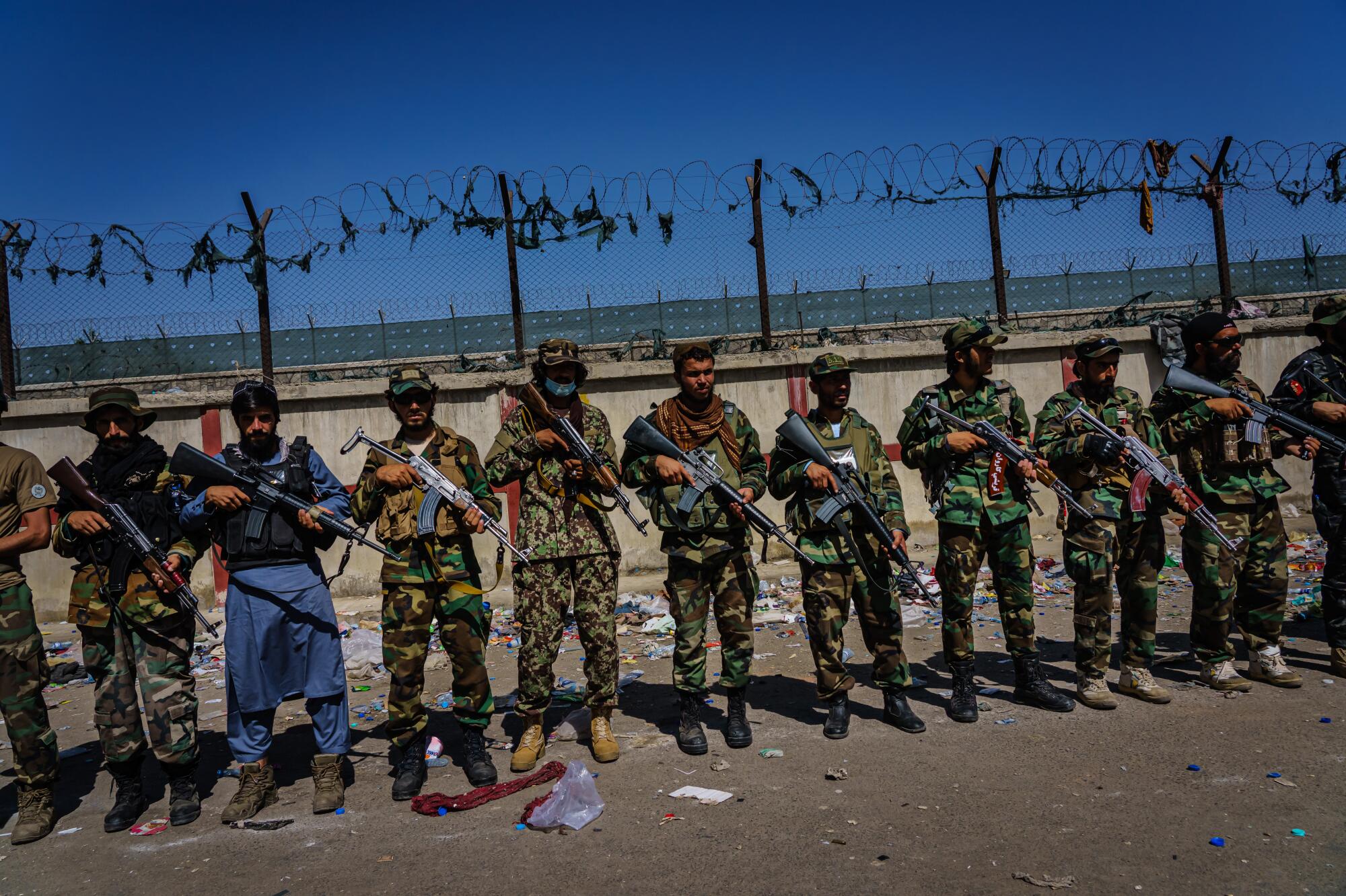 Taliban fighters line up with rifles in front of a fence