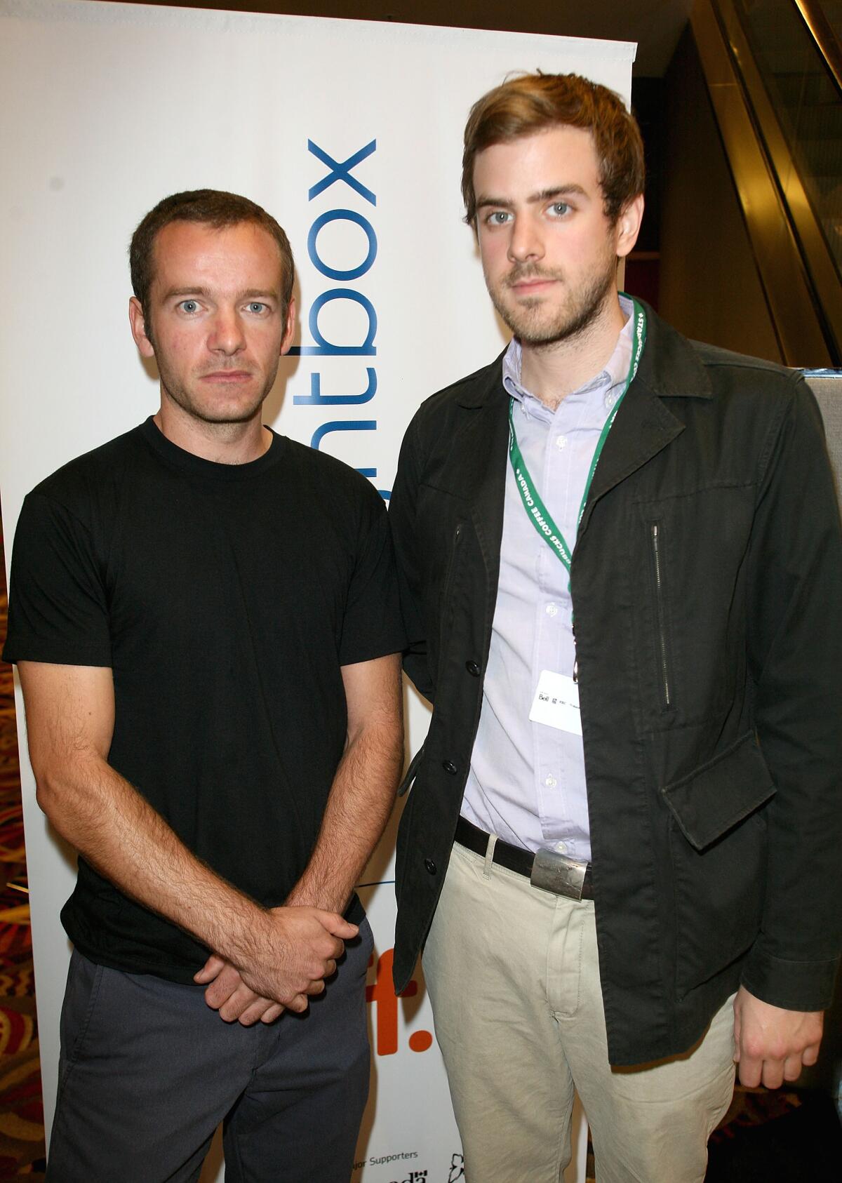 A shorter man clad in a black T-shirt crosses his hands in front of him next to a taller man in a black jacket and khakis