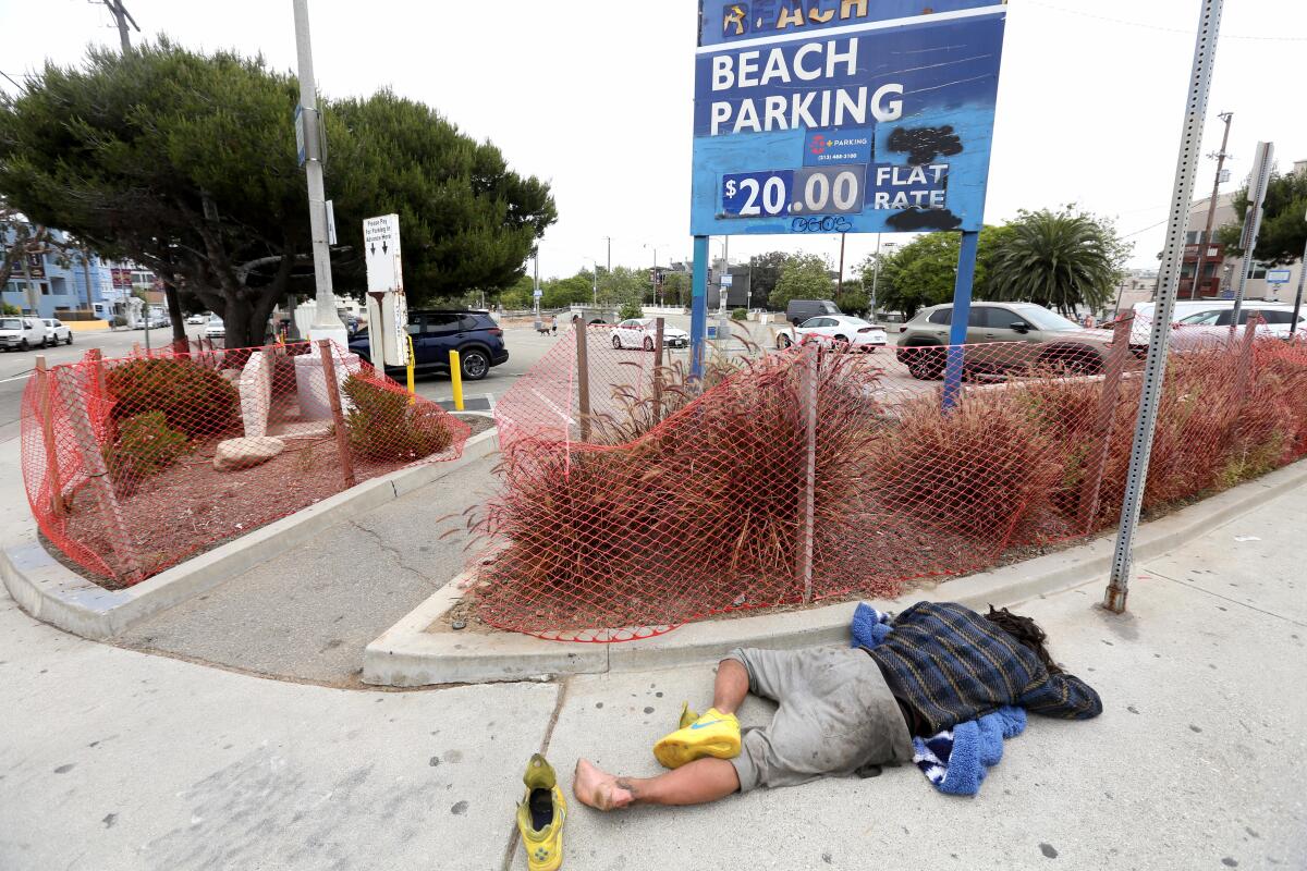 A homeless man sleeps on a sidewalk in front of a parking lot