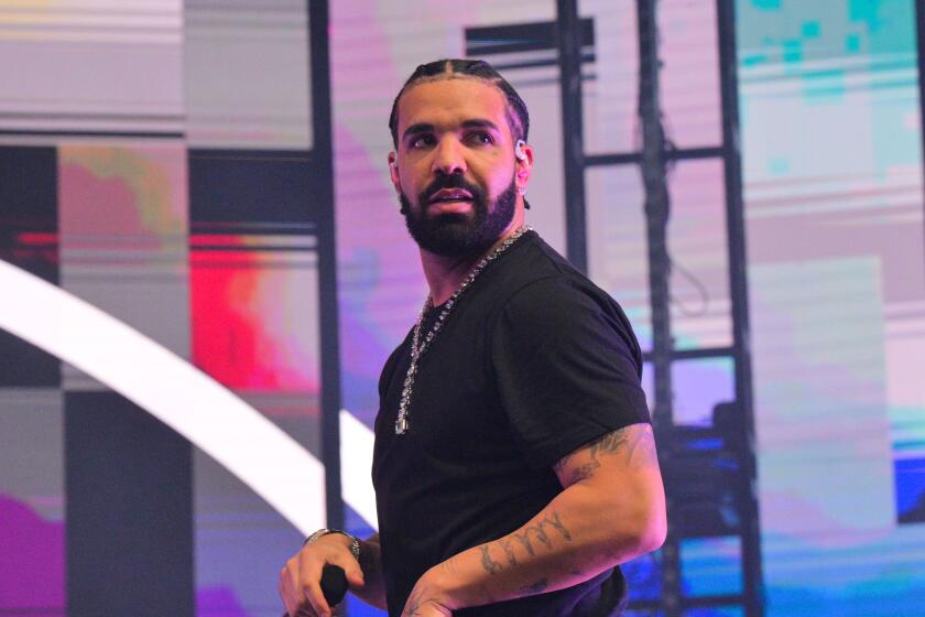 Man, drake, wearing black shirt and diamond chain, walking sideways with hands to side while performing on stage