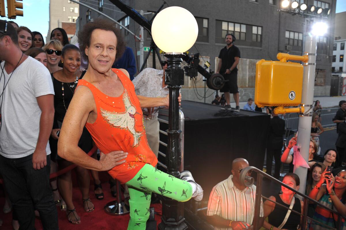 Richard Simmons in a bright orange tank top and neon green leggings holding on to a lamp post.