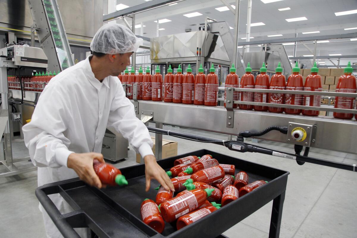 A worker in coveralls and a hairnet grabs bottles of hot sauce from a conveyor belt
