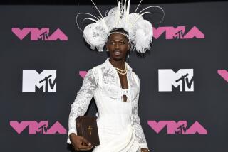 Lil Nas X poses in a white corset and feathered headdress holding a bible next to his right hip