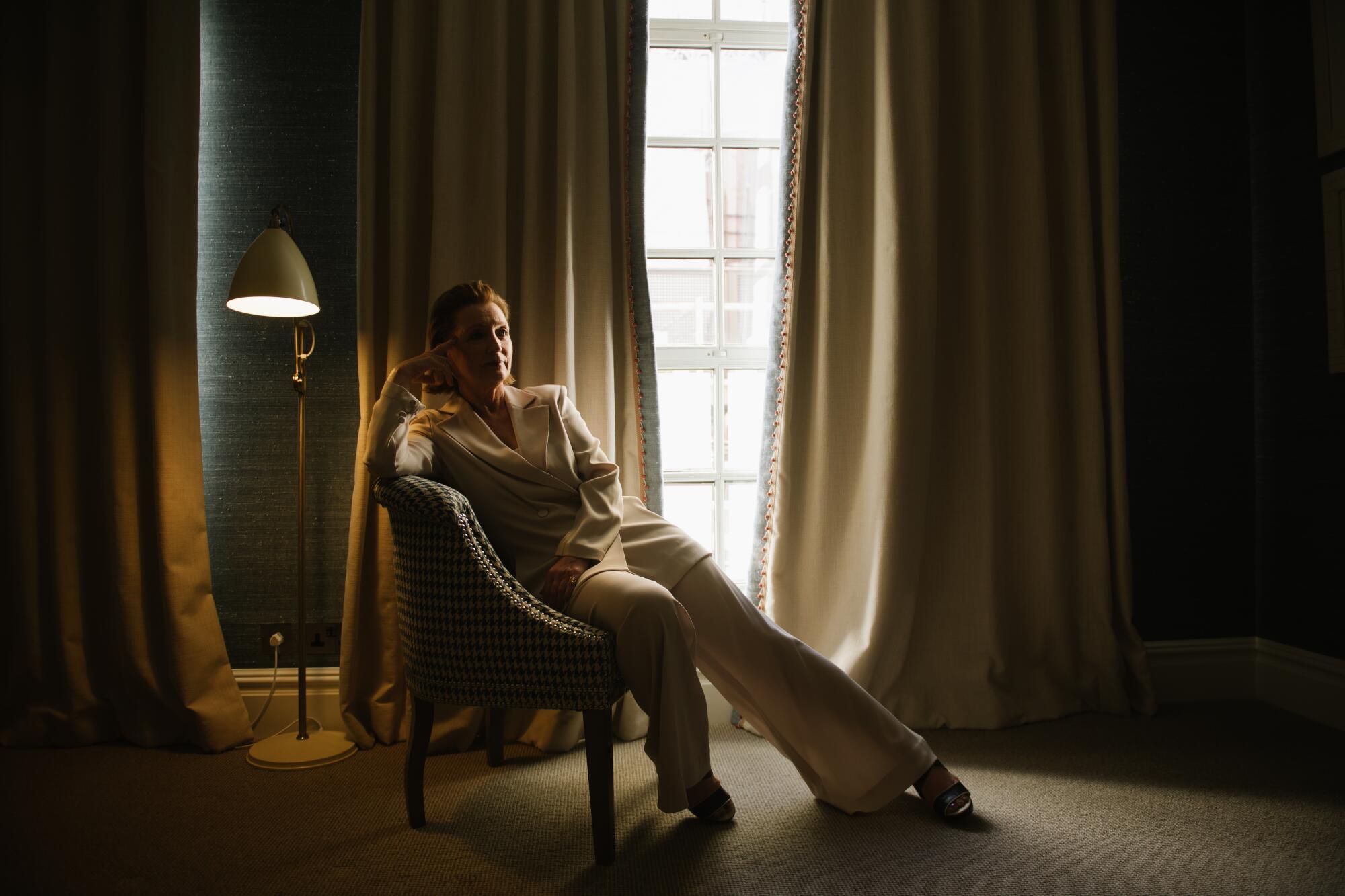 A woman in a white suit sits in a dark room with fancy decor and white curtains