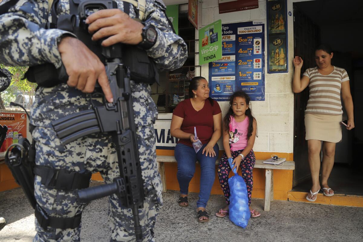 Varias personas sentadas en la entrada de una tienda custodiada por un militar