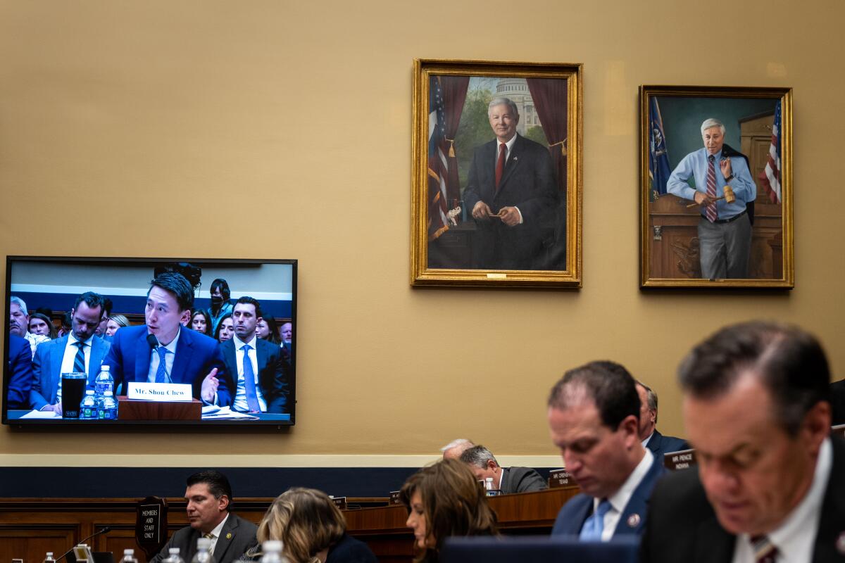 Two portraits hang on a wall next to a screen displaying attendees at a hearing, with people seated in front of the screen