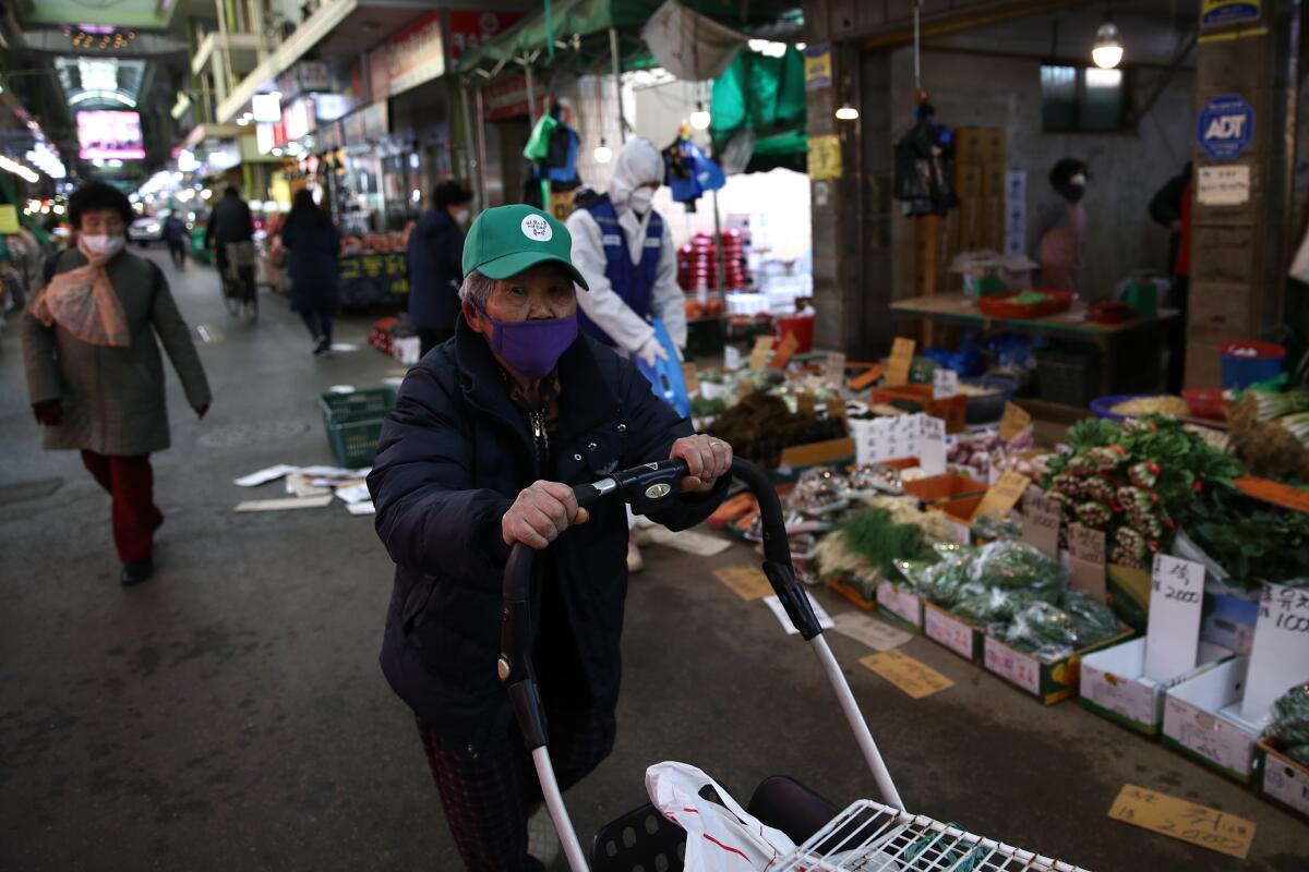 An elderly South Korean man wearing a ball cap and face masks walks in a Seoul market, pushing a cart.