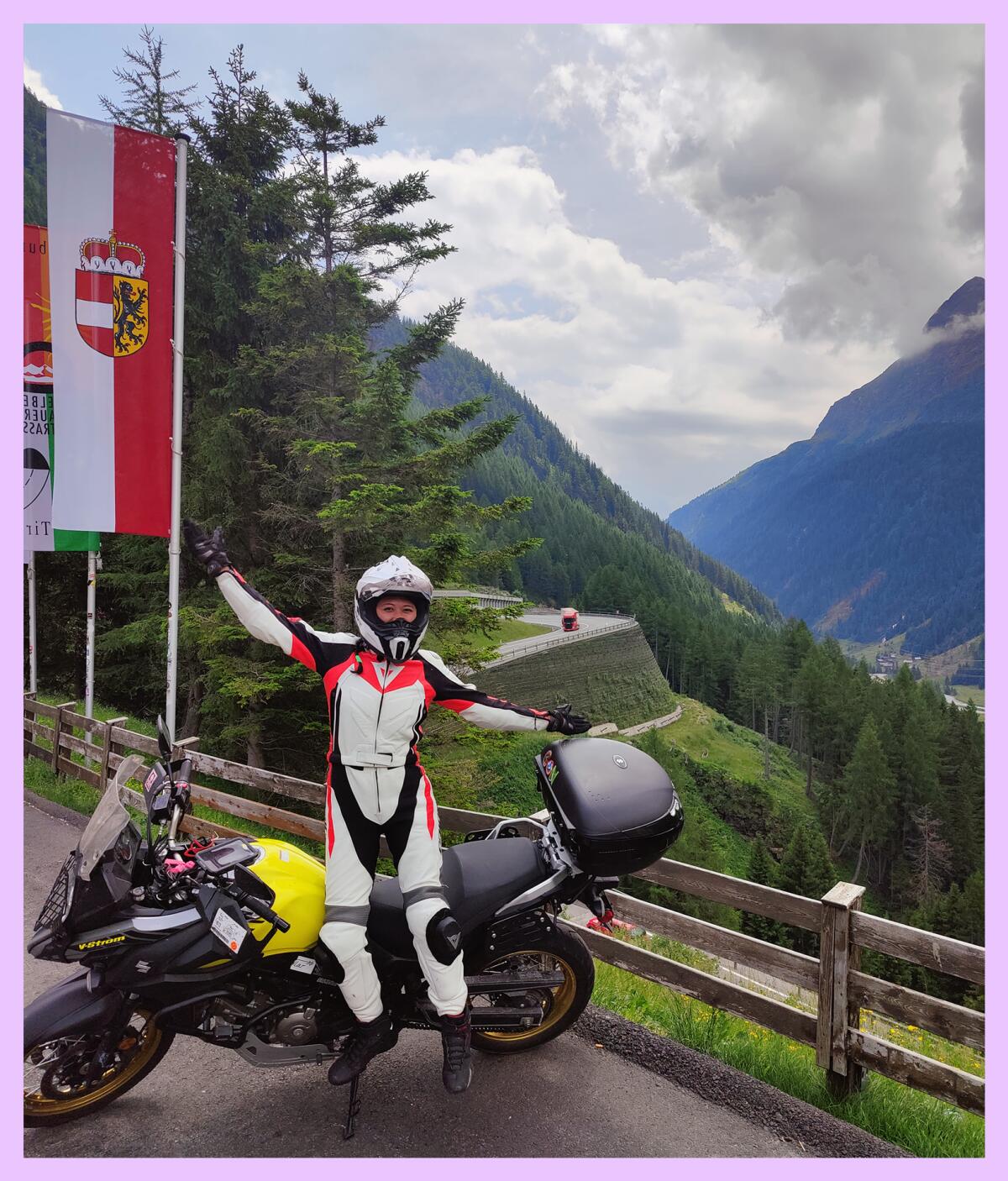 A helmeted person with outstretched arms next to a motorcycle in front of a mountain vista