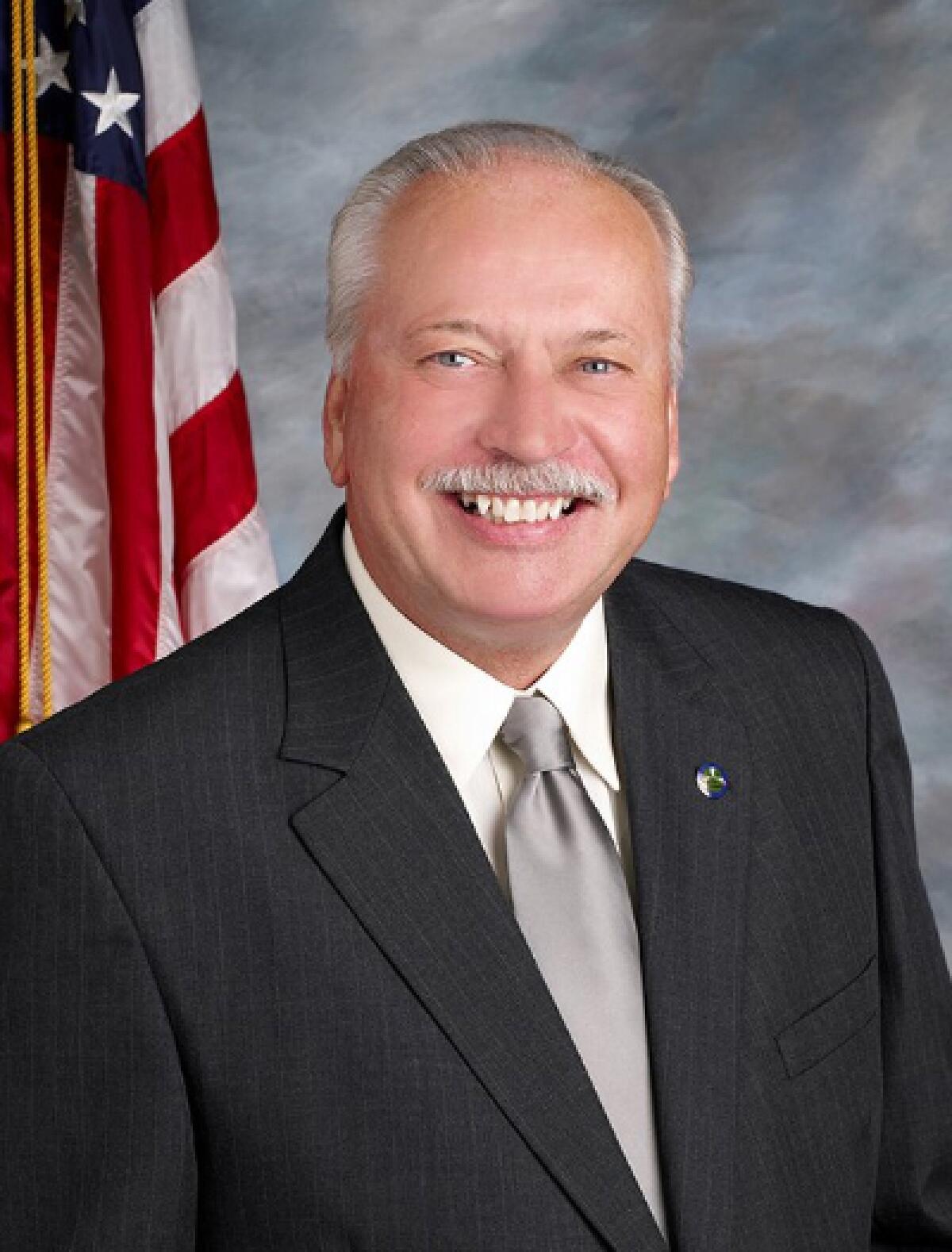 Jim Bowman smiling in a suit with an American flag in the background.