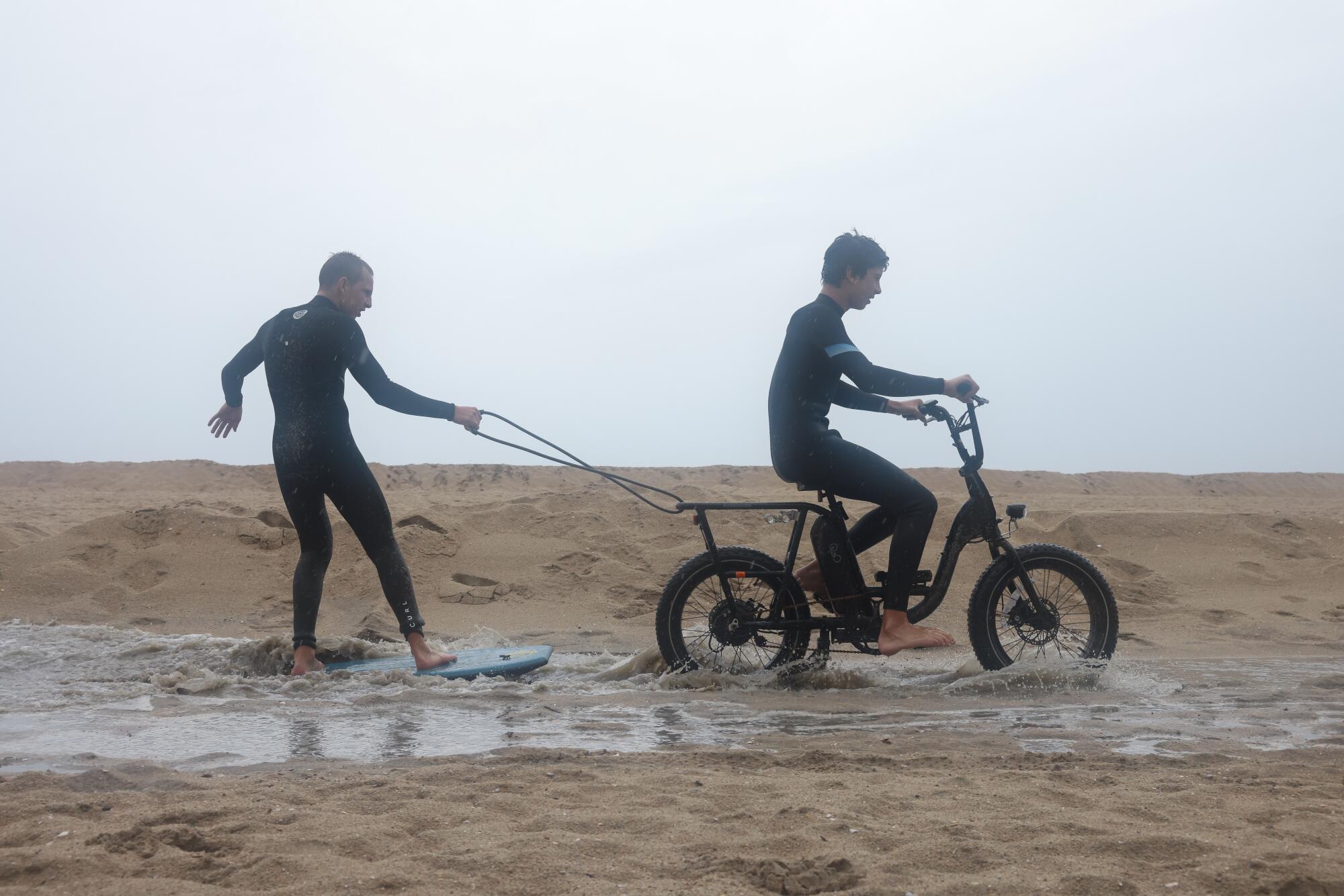 A person on a board holds onto a rope connected to the back of a bicycle that another person is riding